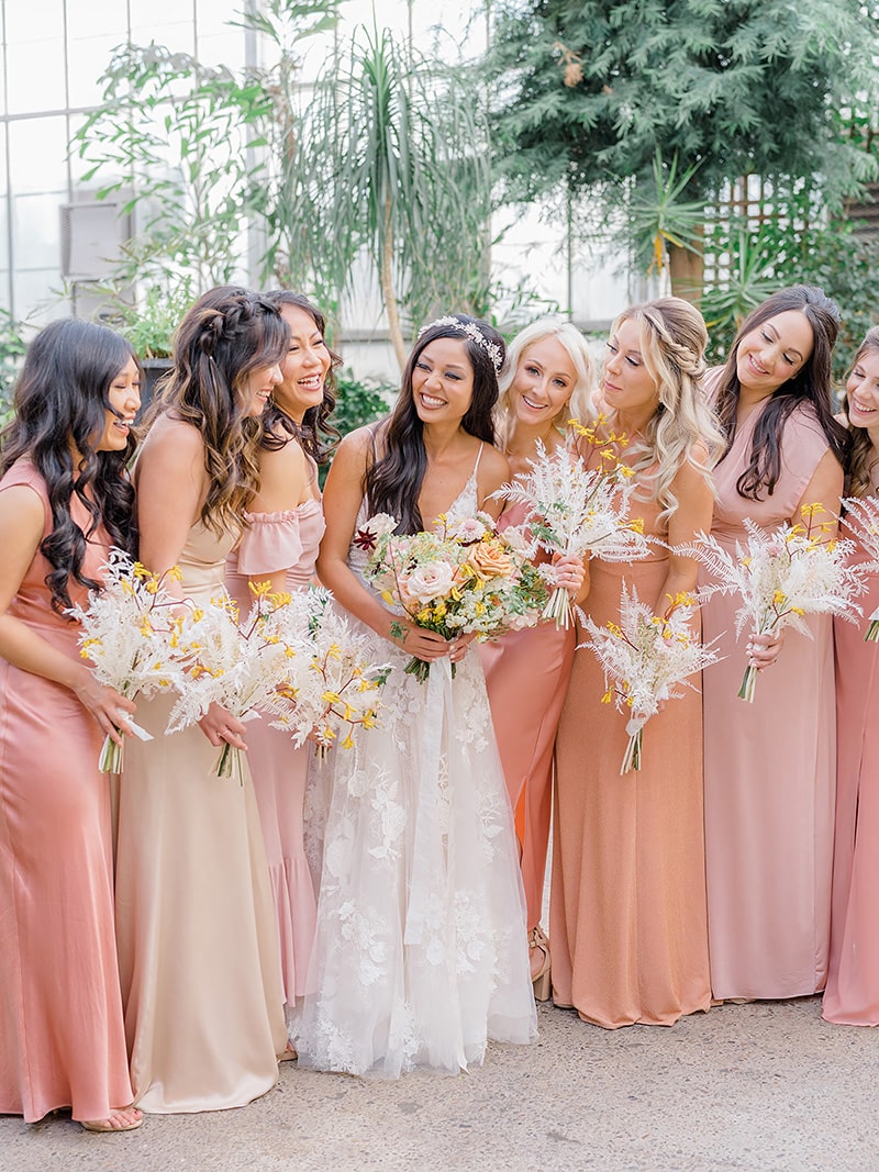 Romantic Boho Desert Pastel Wedding Ideas | Floor length mismatched romantic boho bridesmaid dresses in pink, peach and ivory tones. The wedding party wear their hair in crown braids and loose waves and carry sprays of white dried ruscus and fern paired with yellow kangaroo paw. The bride in the centre of the group wears a tulle wedding dress with spaghetti straps and floral lace applique, a white beaded circlet headpiece and boho waves. She carries a larger bouquet with added roses and other delicate blooms. | Photography: Magdalena Studios via Belovely Floral Co.