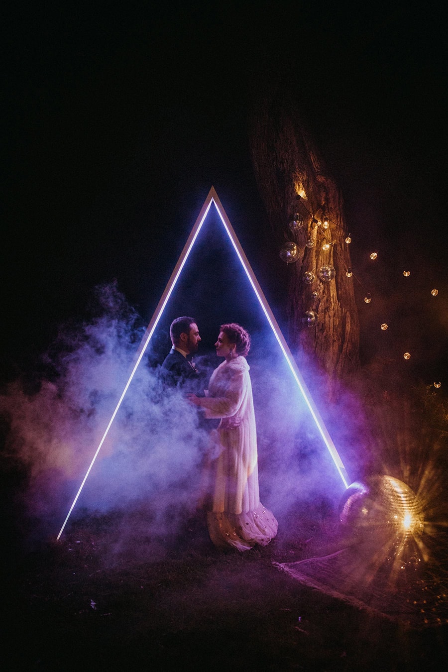 Electric Disco Wedding Ideas | A newlywed couple stands beneath a purple neon triangle wedding ceremony arch at night surrounded by white haze from a smoke bomb that is illuminated by the neon lighting. A large mirror ball is sitting on the ground at the base of the arch reflecting gold light. | Photography: Anna Taylor via Nouba