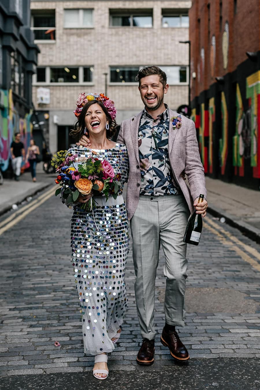 Electric Disco Wedding Ideas |  A bride and groom laugh as they walk down an urban industrial cobbled street with graffic art on the walls. The bride wears a shimmering silver sequin wedding dress. She carries a bright multicolour wedding bouquet and wears a matching flower crown. The groom wears grey pants, muted pink jacket and a tropical print shirt. He carries an open bottle of champagne. | Photography: Epic Love Story