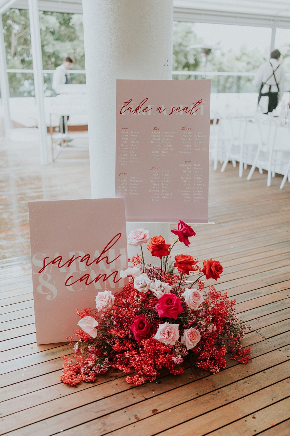 Modern Red & Pink Wedding Ideas | Wedding reception seating chart displayed on an easel features red flowing script layered over bold white lettering on a pink backdrop which reads "Take a Seat". Table numbers and guests are listed below. A second sign displayed below reads "Sarah & Cam" in the same fonts and colours. A modern floral arrangement consisting of a cloud of red painted baby's breath, and sprouting red and pink roses completes the display. | Photography: Alana Taylor Photography via Hello May