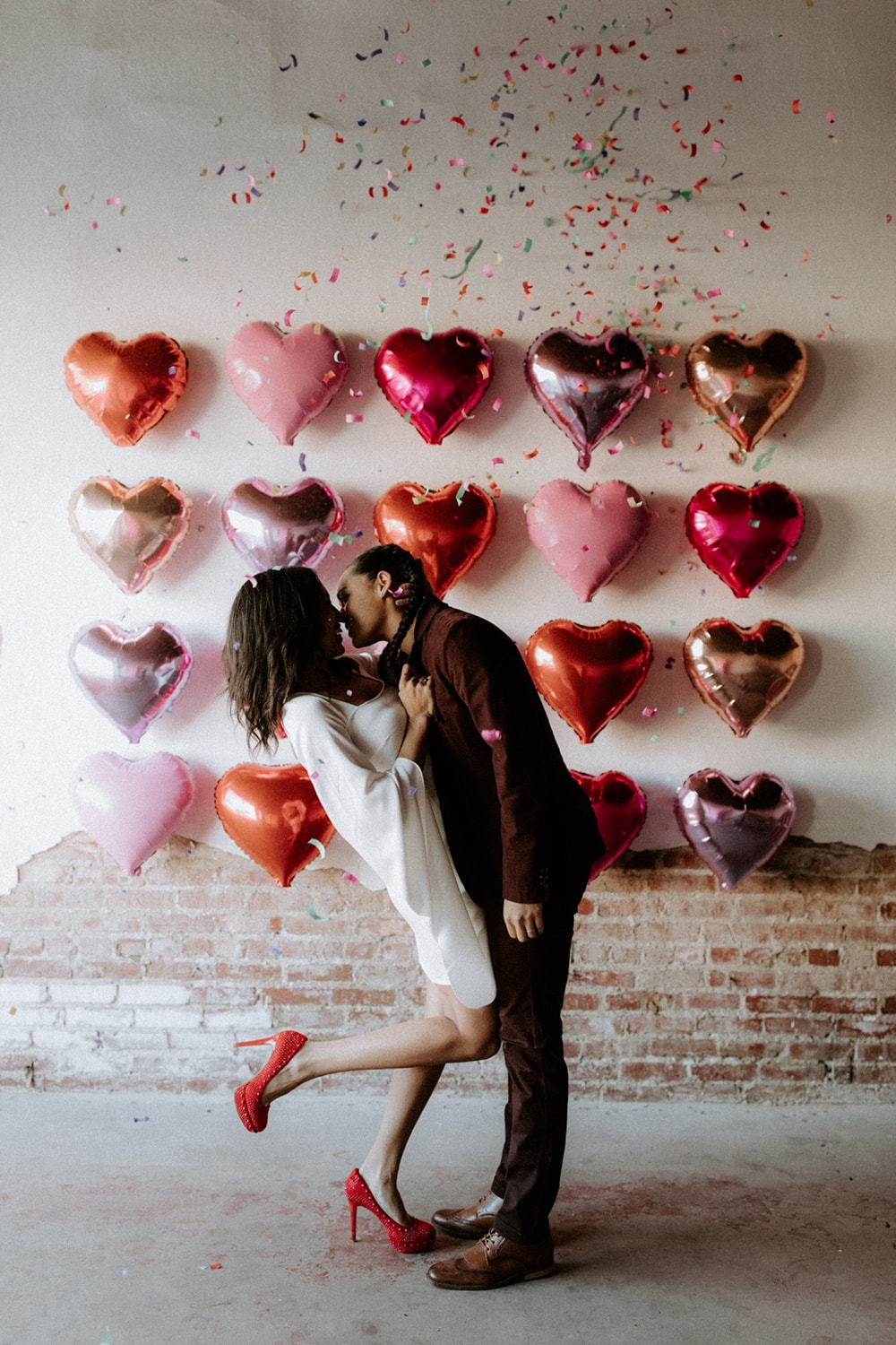 Modern Red & Pink Wedding Ideas | A fun wedding photo backdrop on an industrial brick wall consisting of four rows of metallic foil heart balloons in shades of pink, red and orange. A same-sex couple kisses in front of the backdrop as rainbow confetti rains down on them. One wears a short white dress with long flowing sleeves and red heels. The other wears a burgundy suit with brown leather shoes. | Photography: The Dogwood Collective via Junebug Weddings