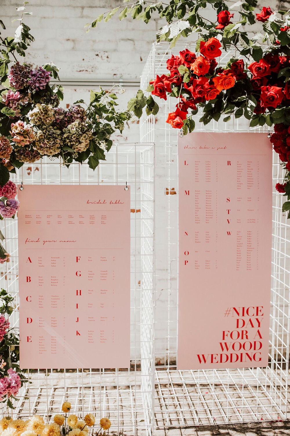 Modern Red & Pink Wedding Ideas | A modern wedding reception seating chart featuring red lettering on a blush pink backdrop attached to a white rectangular metal pillar that is decorated with red roses and greenery. The text reads "Nice Day for a Wood Wedding" | Photography: Casie Marie Photography via Ruffled