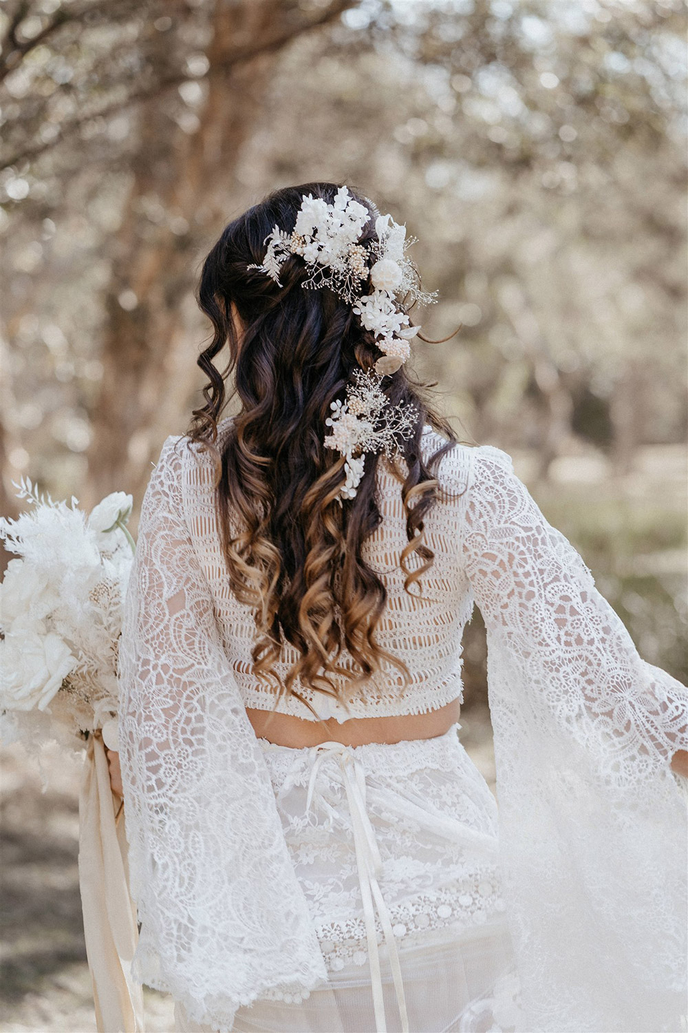 Outdoor Festival Bohemian Wedding Inspiration | The bride wears a white two piece lace boho wedding dress with a long sleeved cropped bodice and tiered maxi skirt. Her hair is worn in a romantic half up half down style with loose waves and a cascading fresh floral hairpiece. | Photography: Shae Estella Photo