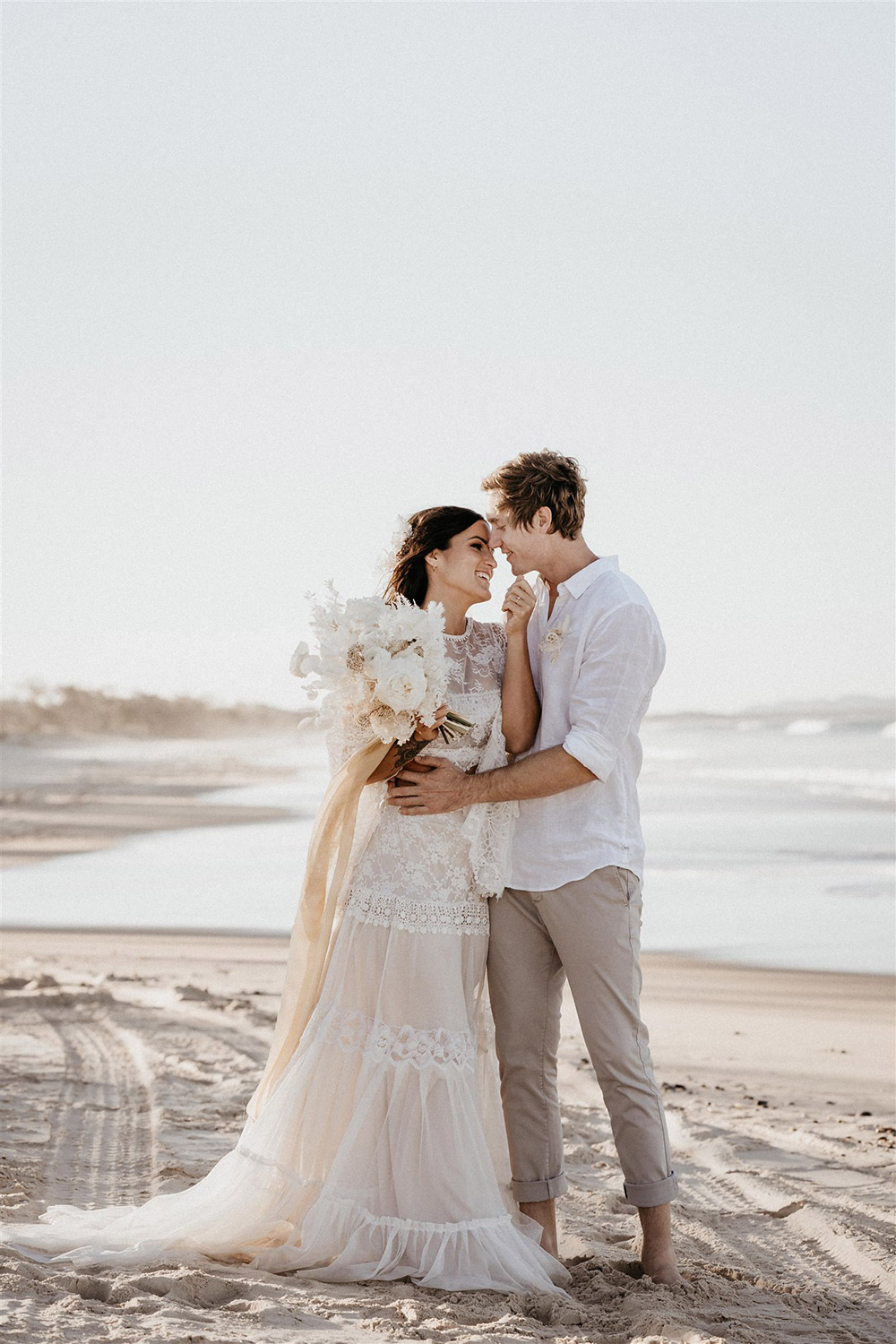 Outdoor Festival Bohemian Wedding Inspiration | The bride wears a white two piece lace boho wedding dress with a long sleeved cropped bodice and tiered maxi skirt. Her hair is worn in a romantic half up half down style with loose waves and a cascading fresh floral hairpiece. The groom wears an untucked white linen shirt with the sleeves rolled up and collar unbuttoned, light grey chinos and brown boots for a casual wedding day look. | Photography: Shae Estella Photo