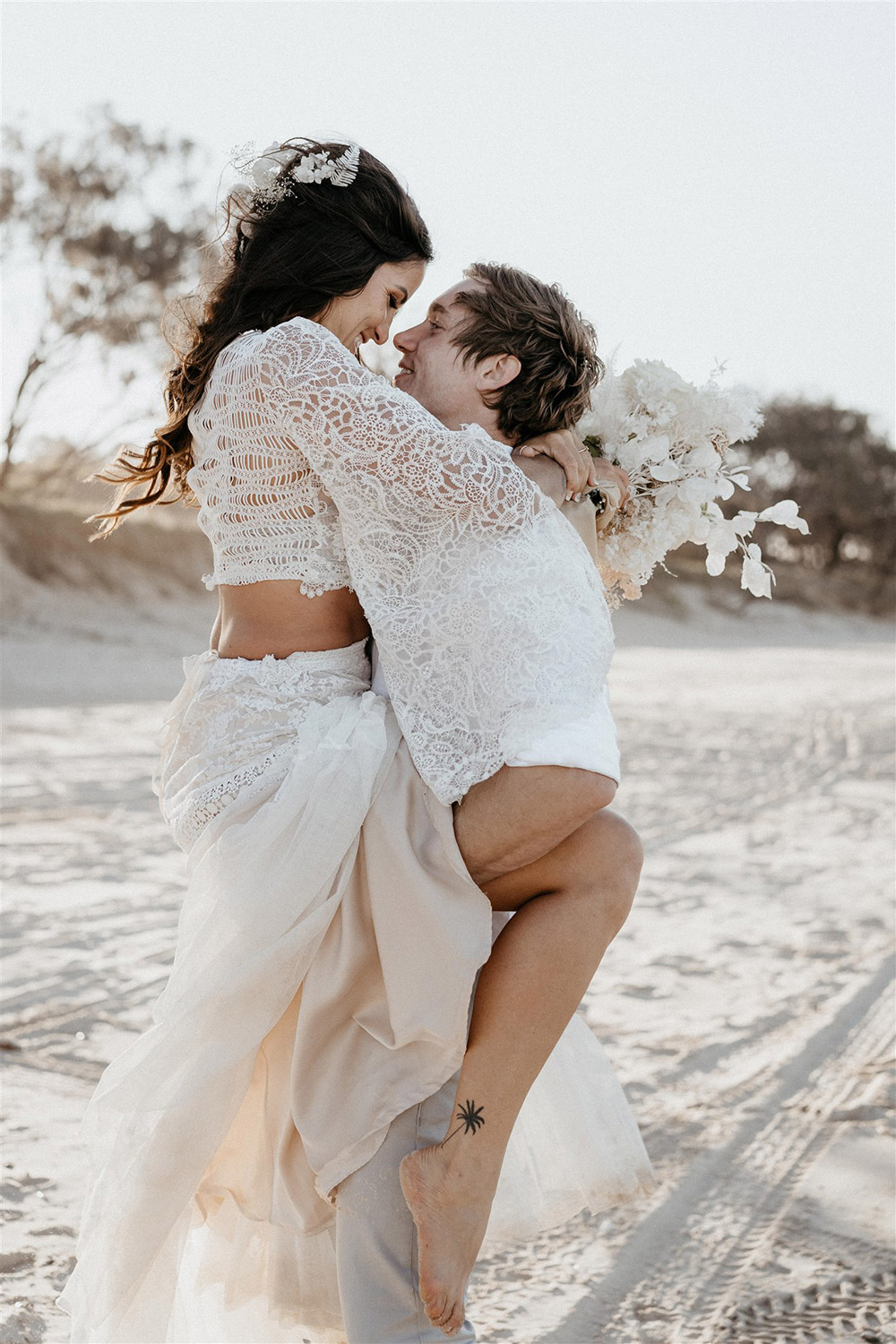 Outdoor Festival Bohemian Wedding Inspiration | The bride wears a white two piece lace boho wedding dress with a long sleeved cropped bodice and tiered maxi skirt. Her hair is worn in a romantic half up half down style with loose waves and a cascading fresh floral hairpiece. The groom wears an untucked white linen shirt with the sleeves rolled up and collar unbuttoned, light grey chinos and brown boots for a casual wedding day look. | Photography: Shae Estella Photo