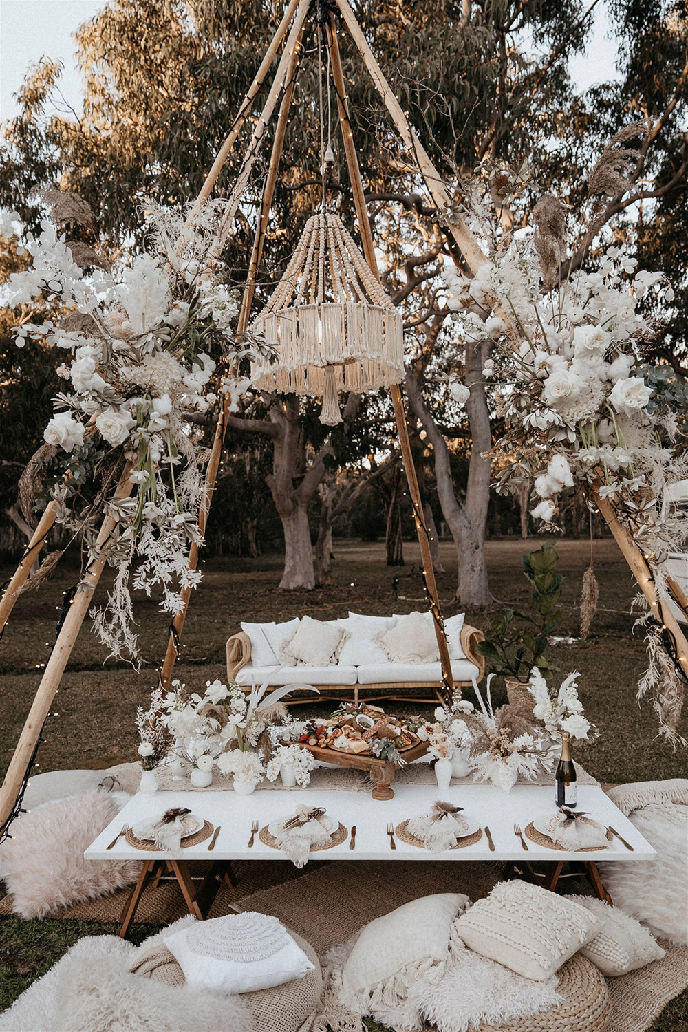 Outdoor Festival Bohemian Wedding Inspiration | A naked tipi wrapped in fairy lights and sprays of boho florals is set for picnic style grazing. Beneath a beaded and fringed chandelier sits layered rugs, scattered cushions in neutral and white textures and a low table set for eight. This is topped with a macrame runner and clusters of white vessels displaying fresh posies. The place settings feature gold cutlery, a natural woven placemat topped with white plateware and neutral napkins tied with cord and feathers. A generous antipasto platter placed in the centre of the table is ready for the happy couple and their crew! | Photography: Shae Estella Photo
