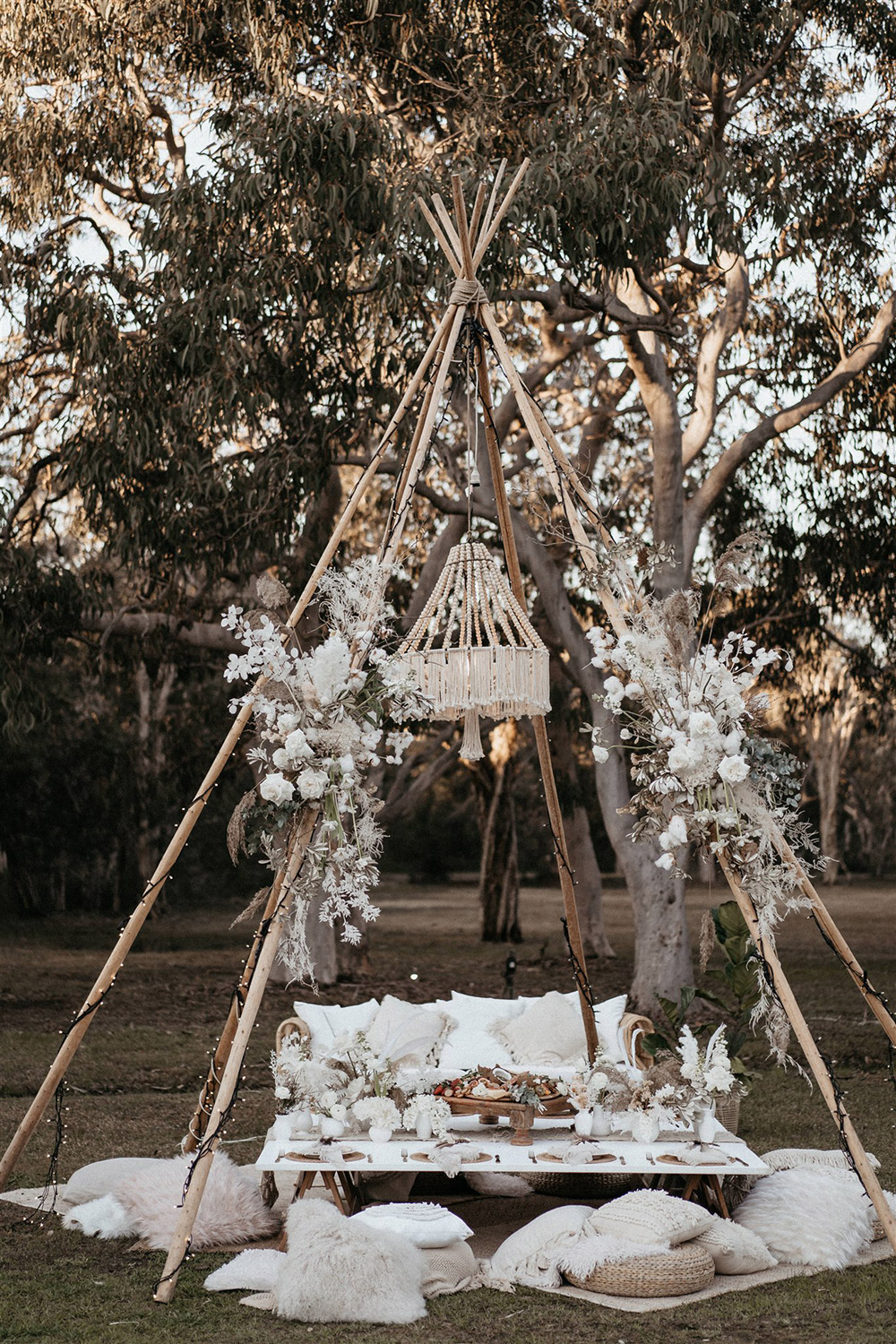 Outdoor Festival Bohemian Wedding Inspiration | A naked tipi wrapped in fairy lights and sprays of boho florals is set for picnic style grazing. Beneath a beaded and fringed chandelier sits layered rugs, scattered cushions in neutral and white textures and a low table set for eight. This is topped with a macrame runner and clusters of white vessels displaying fresh posies. | Photography: Shae Estella Photo