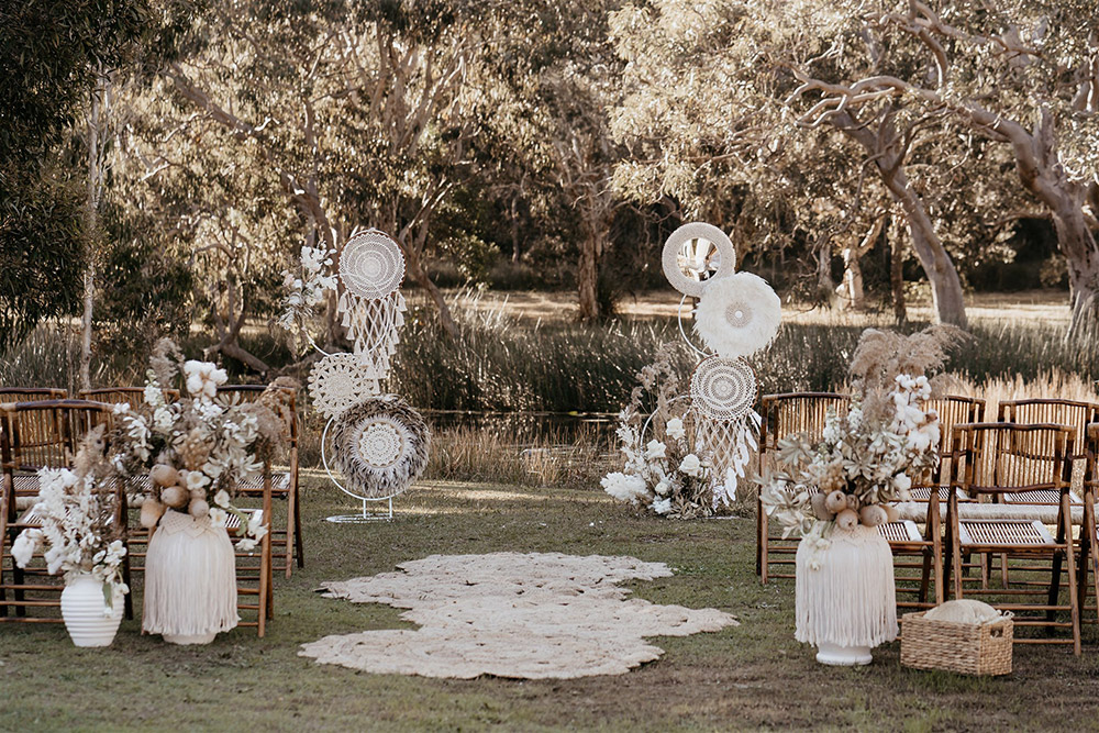 Outdoor Festival Bohemian Wedding Inspiration | This outdoor boho wedding ceremony consists of woven jute rugs for the aisle and two pillars of white circles in varying sizes adorned with macrame, feathers and flowers for the arbour. The setting is framed by bamboo folding chairs for guests and large white vases decorated with fringed macrame wraps that are filled with neutral dried floral arrangements accented with stems of white tulips, stocks, cotton and ruscus. | Photography: Shae Estella Photo