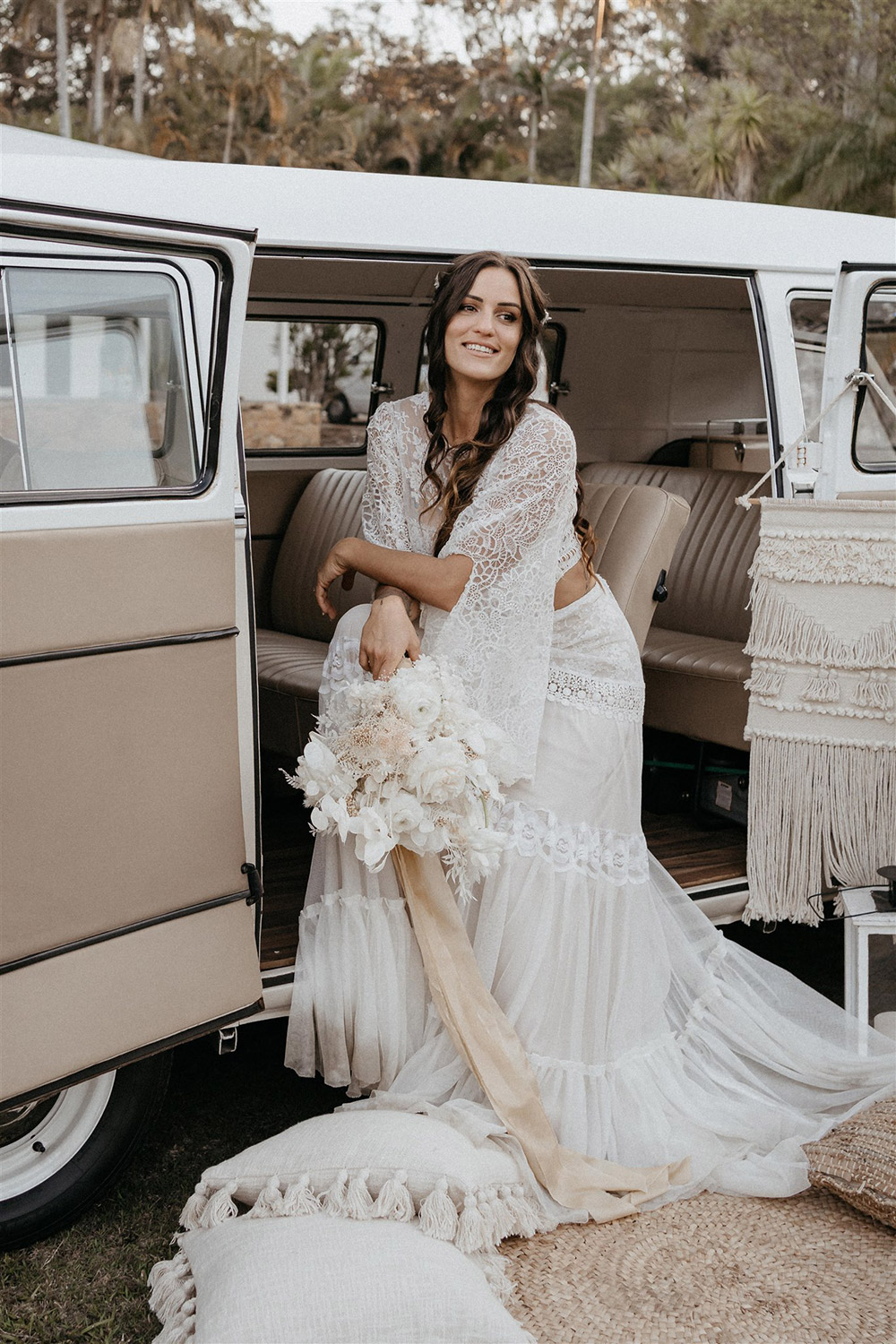 Outdoor Festival Bohemian Wedding Inspiration | Sitting in a neutral coloured Kombi, the bride wears a white two piece lace boho wedding dress with a long sleeved cropped bodice and tiered maxi skirt. Her hair is worn in a romantic half up half down style with loose waves and a cascading fresh floral hairpiece. | Photography: Shae Estella Photo
