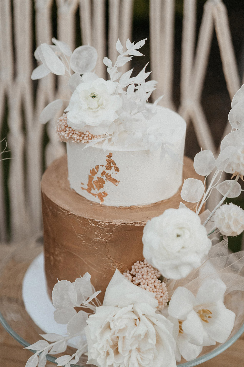 Outdoor Festival Bohemian Wedding Inspiration | A two tiered wedding cake sits on a clear glass cake plate. The top tier features smooth white buttercream with a trail of gold leaf, while the bottom tier features textured gold buttercream. The cake is decorated with artful sprays of white dried silver dollar eucalyptus, ruscus, scabiosa pods and skeleton leaves, blush dried rice flower and fresh white roses, tulips and ranunculus. | Photography: Shae Estella Photo