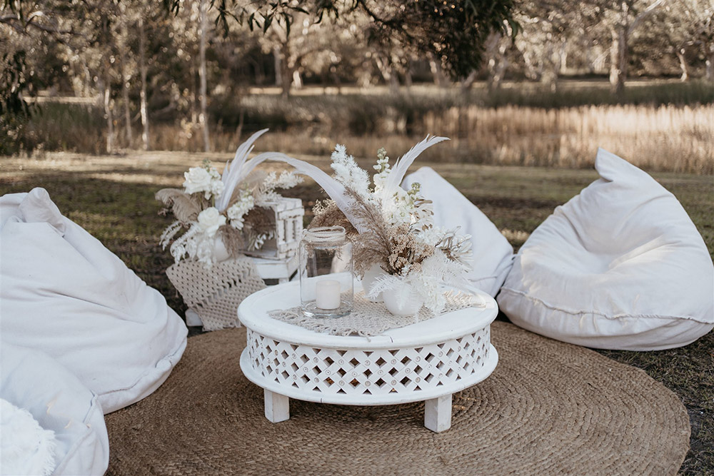 Outdoor Festival Bohemian Wedding Inspiration | The outdoor cocktail style wedding reception features a number of eclectic seating areas for guests. Here, white bean bags encircle a natural round jute rug. A small white round wooden coffee table and rustic white stacked crates complete the setting, which are decorated with macrame runners, a pillar candle in a clear glass hurricane lantern and small vases of white and neutral flowers, feathers and foliage. | Photography: Shae Estella Photo