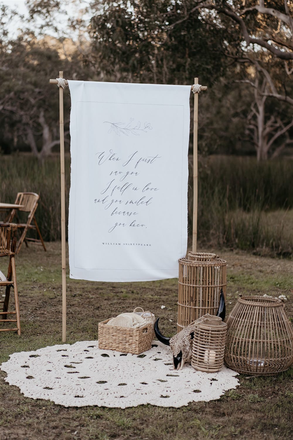 Outdoor Festival Bohemian Wedding Inspiration | A white fabric wedding welcome sign features the same fine line foliage illustration as the stationery suite and the William Shakespeare quote, "When I first saw you I fell in love and you smiled because you knew." A cluster of bamboo lanterns and a woven basket of jute fans sits atop a macrame rug alongside the welcome sign. | Photography: Shae Estella Photo
