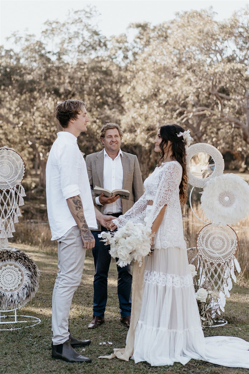 Outdoor Festival Bohemian Wedding Inspiration | The bride wears a white two piece lace boho wedding dress with a long sleeved cropped bodice and tiered maxi skirt. Her hair is worn in a romantic half up half down style with loose waves and a cascading fresh floral hairpiece. The groom wears an untucked white linen shirt with the sleeves rolled up and collar unbuttoned, light grey chinos and brown boots for a casual wedding day look. | Photography: Shae Estella Photo