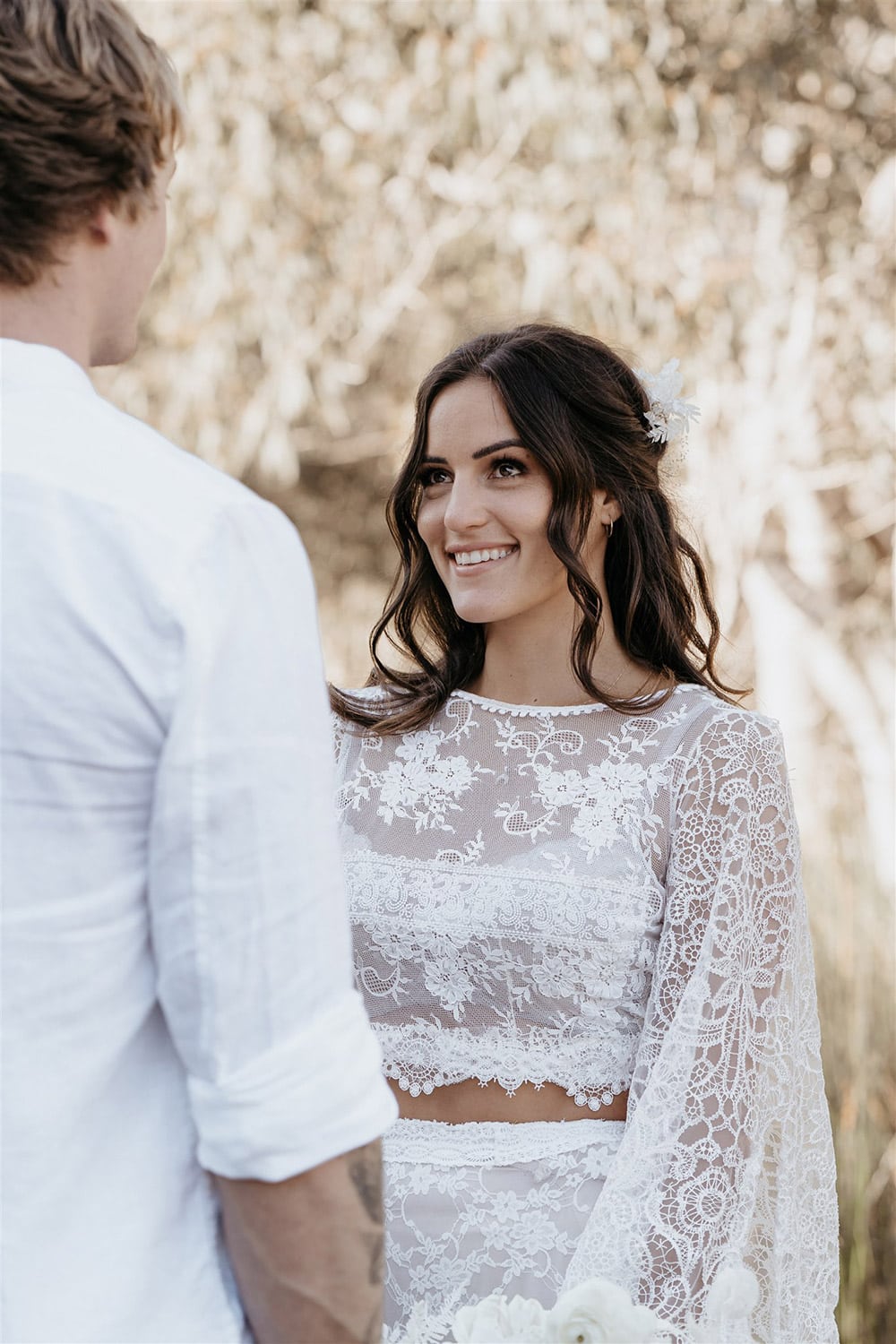 Outdoor Festival Bohemian Wedding Inspiration | The bride wears a white two piece lace boho wedding dress with a long sleeved cropped bodice and tiered maxi skirt. Her hair is worn in a romantic half up half down style with loose waves and a cascading fresh floral hairpiece. | Photography: Shae Estella Photo