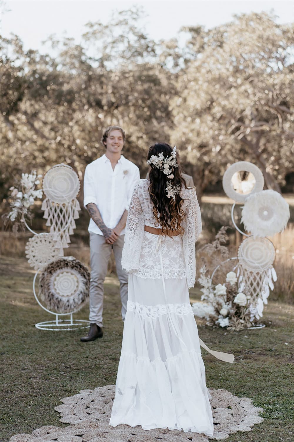 Outdoor Festival Bohemian Wedding Inspiration | The bride wears a white two piece lace boho wedding dress with a long sleeved cropped bodice and tiered maxi skirt. Her hair is worn in a romantic half up half down style with loose waves and a cascading fresh floral hairpiece. The groom wears an untucked white linen shirt with the sleeves rolled up and collar unbuttoned, light grey chinos and brown boots for a casual wedding day look. | Photography: Shae Estella Photo