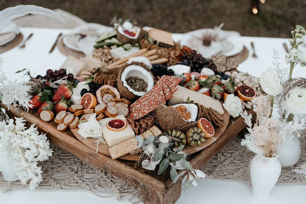 Outdoor Festival Bohemian Wedding Inspiration | A white timber picnic table is topped with a macrame runner, clusters of white vessels displaying fresh posies and a generous antipasto grazing platter ready for the happy couple and their crew! | Photography: Shae Estella Photo