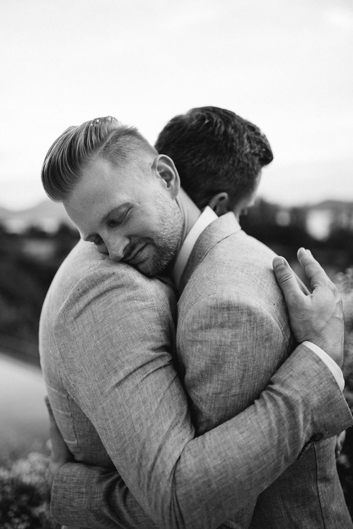 Two grooms wearing suits embrace one another during their wedding portraits, resting their heads on each other's shoulders. | Photography: Alen Karupovic via Junebug Weddings