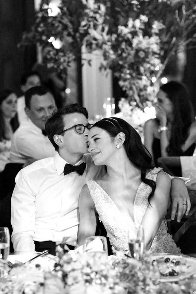 A newly married couple is seated at their reception listening to speeches. The bride leans into the groom as he kisses her on the temple. | Photography: Sarah Falugo via Brides