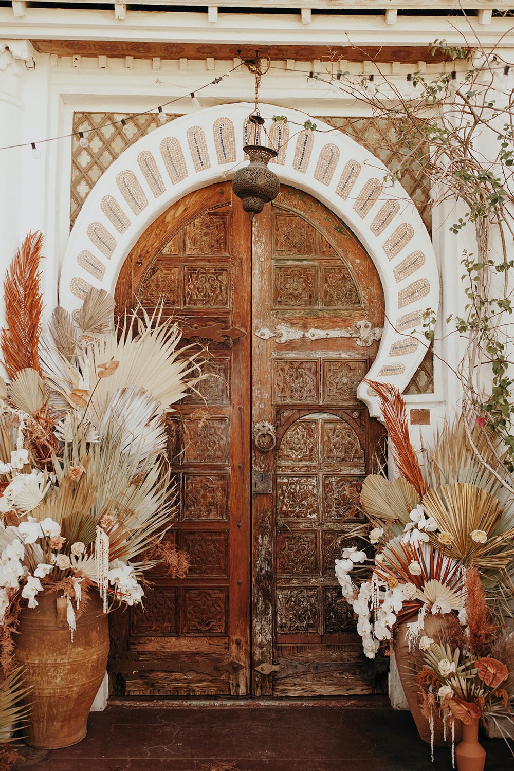 Boho Rust & White Wedding Ideas | An ornately carved wooden door inspired by Moroccan architecture used as a boho wedding ceremony backdrop. It is framed by large ceramic pots filled with wild floral arrangements consisting of dried palm leaves and anthurium in earthy colours, white orchids, amaranthus and roses. | Photography: Jordan Voth via Foxtail Florals