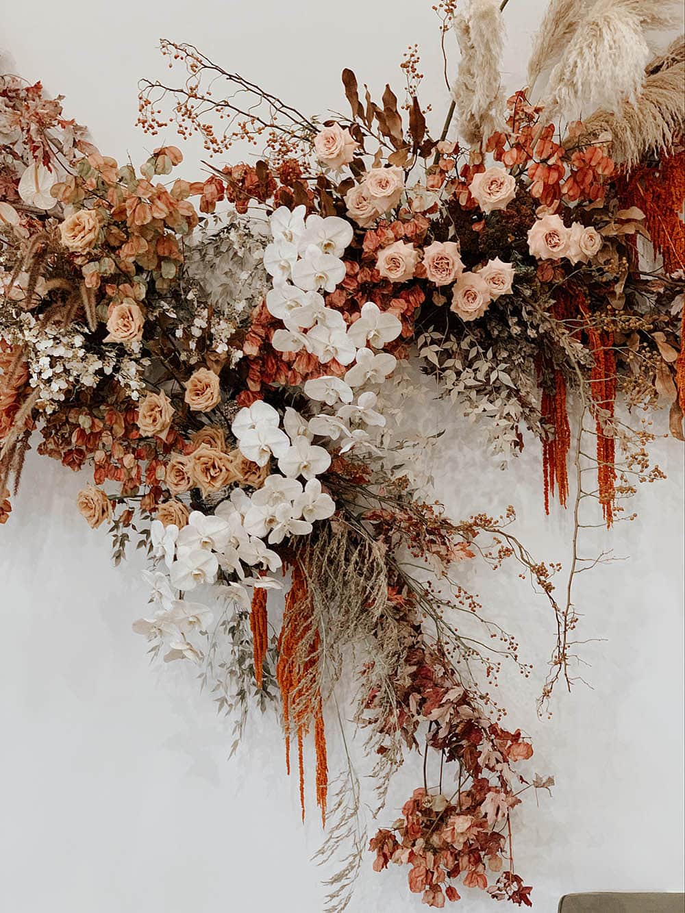 Boho Rust & White Wedding Ideas | An earthy boho wedding ceremony backdrop consisting of a floating asymmetrical floral arrangement hung on a white wall which features blush roses, white orchids, rust coloured bougainvillea and dripping amaranthus, and neutral feathery foliage. | Photography & Flowers: Nikau Flora
