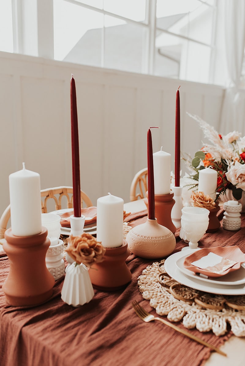 Romantic Boho Terracotta Wedding Ideas | An earthy boho wedding reception table with rattan dining chairs features alternating terracotta and white candles in ceramic candle holders of different shapes and sizes on a terracotta cheesecloth table runner alongside brass cutlery, natural woven placemats and layered white plateware topped with a leaf shaped ceramic dish and torn edge paper place card. | Photography: Bruna Kitchen