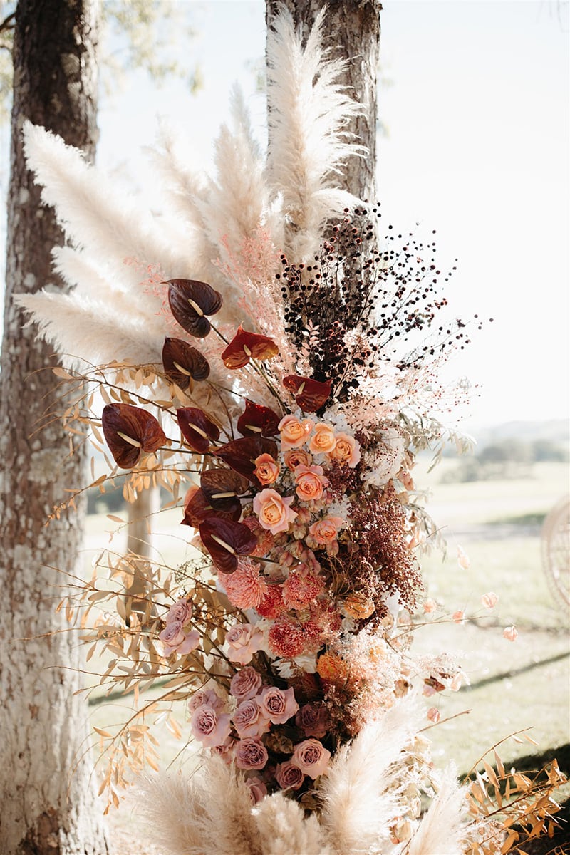 Romantic Boho Terracotta Wedding Ideas | Wild boho wedding ceremony floral pillar featuring merlot anthurium, peach and lilac roses, pink chrysanthemum disbuds and sprays of purple stirlingia, pale pink ruscus, neutral pampas grass and dried foliage. | Photography: Carly Tia via Hello May