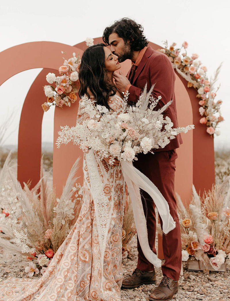 Romantic Boho Terracotta Wedding Ideas | A trio of terracotta and clay wedding ceremony arches in a desert landscape decorated with neutral, peach, pink and white flowers. A couple stands in front of the ceremony backdrop. The bride wears a terracotta coloured wedding dress with bold lace detail holding a bouquet featuring a spray of blush and white flowers and trailing white ribbons. The groom wears a dark terracotta suit and tie with dusty orange shirt and brown leather shoes as he caresses his bride's face and kisses her on the cheek. | Photography: Melissa Marshall via Green Wedding Shoes