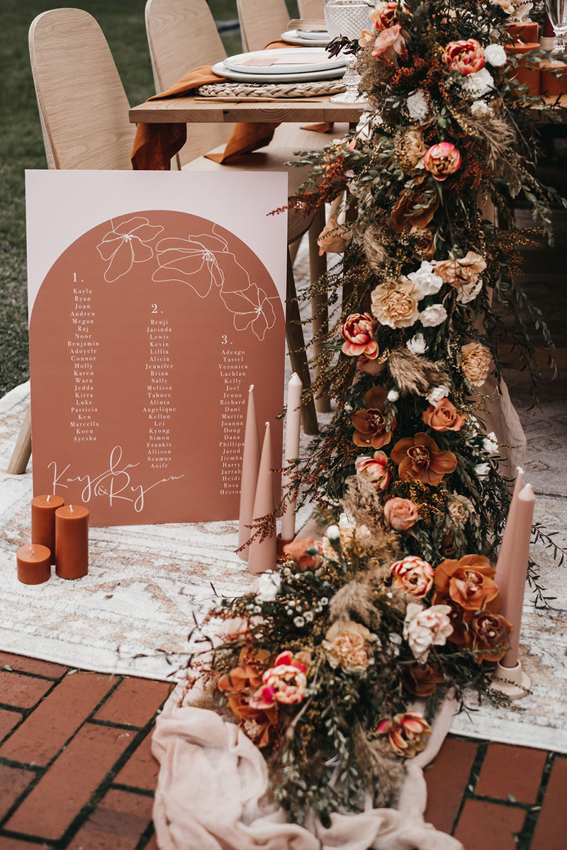 Romantic Boho Terracotta Wedding Ideas | Boho wedding reception table styling with a lush floral garland featuring tulips, orchids, roses and carnations in earthy tones that cascades onto the floor alongside clusters of clay and rust coloured pillar candles and an arch shaped terracotta seating chart with fine line floral illustrations and guest names in white ink. | Photography: Will and Co via Wedshed