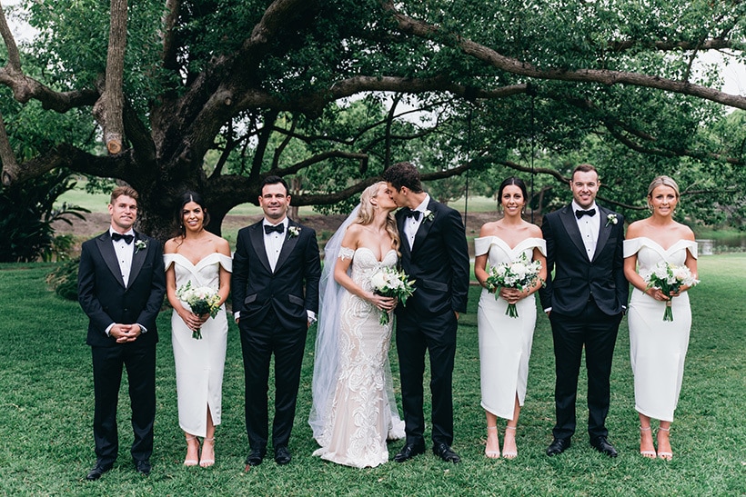 Modern bridal party with groomsmen wearing black suits, bow ties and white rose buttonholes with bridesmaids wearing white off the shoulder cocktail dresses | Figtree Wedding Photography