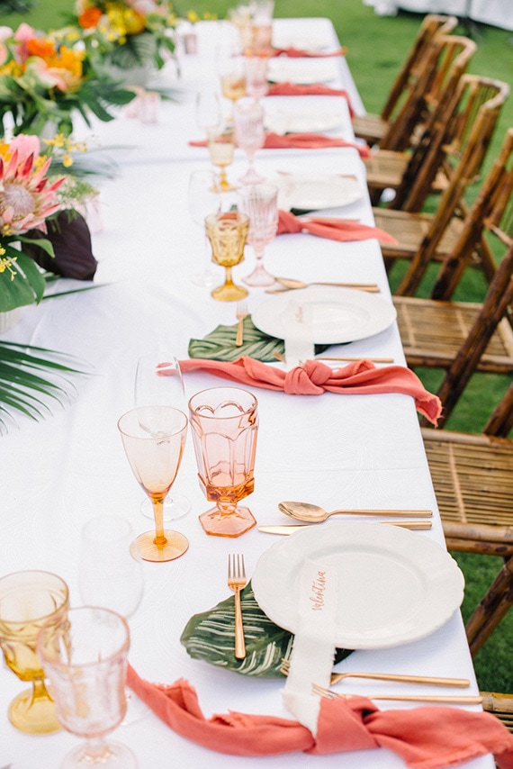 Tropical wedding reception place settings with peach and yellow coloured glassware, palm leaves, gold cutlery and bright coral napkins | Krista Mason Photography via Ruffled